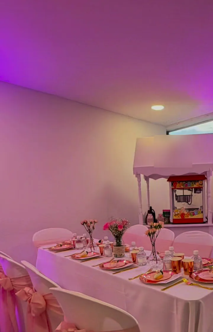Mesa con mantel blanco, sillas con lazos rosados y carrito de crispetas bajo luz rosada